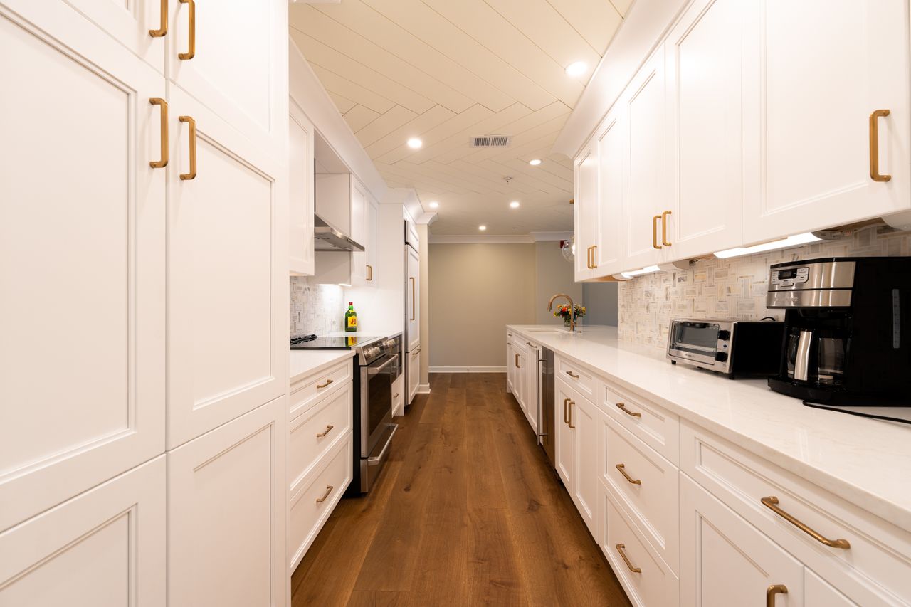 gallery-4 Bright modern kitchen with white cabinets, wooden floor, and appliances.